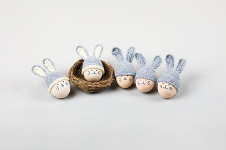 Five Easter eggs with funny faces and gray crocheted hats with bunny ears in a decorative nest of...