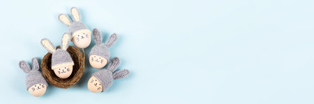 Five Easter eggs with funny faces and gray crocheted hats with bunny ears in a decorative nest of sisal on a blue background. Happy Easter concept. Bannerの写真素材