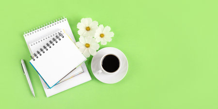 Spring composition with opened blank notepad and pen, cup of black coffee, flowers of a white cosmos on a green background. Festive office desktop concept. Morning coffee cup.の写真素材