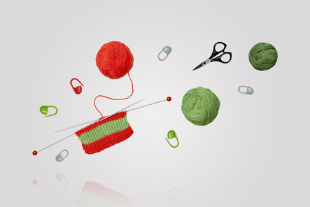 Tools for knitting warm clothes: scissors, green and red balls of thread, stoppers, and knitting needles with knitted green red piece of fabric flying in air on gray background. Hobby home crafts.の写真素材