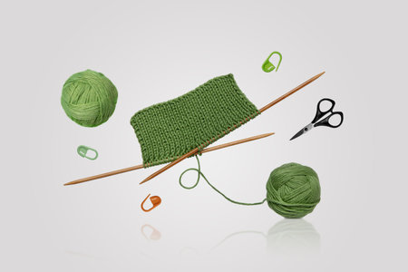 Tools for knitting warm clothes: scissors, green ball of thread, stoppers, and knitting needles with a knitted green piece of fabric on a gray background. Hobby home crafts.の写真素材