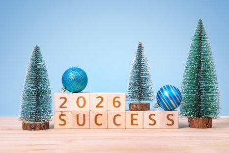 New Year 2026 and Christmas postcard. Wooden cubes with numbers 2026 and word success and green Christmas trees on a wooden table and a blue background.の写真素材