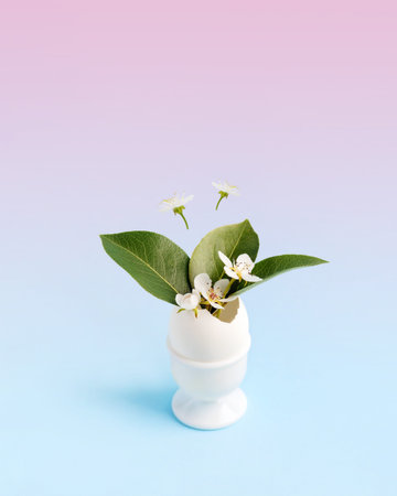 White Easter egg in a stand with spring pear flowers and leaves on a blue pink gradient background...
