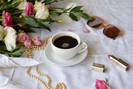 White cup of black coffee on a plate surrounded white pink flowers, sunglasses, lipstick and pearl necklaceの写真素材