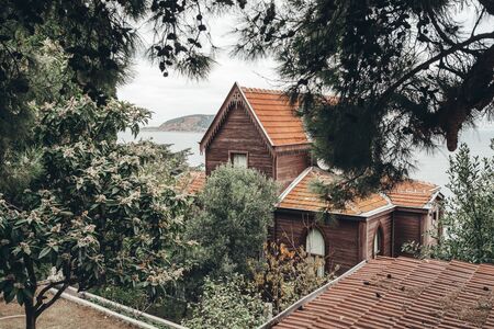 Old brown vintage wooden house with a red tiled roof and beautiful windows in the thickets of green vegetation on the seashore. Sea landscape with a house on a background of mountains and greeneryの写真素材