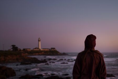 A girl looks at a ship lighthouse with buildings on a rocky coast against the backdrop of the Pacific Ocean with waves and a purple evening sky on a beach in Californiaの写真素材