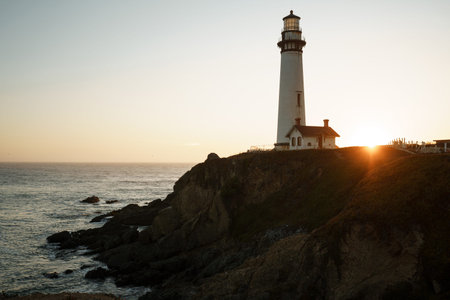 Sunset on the ocean against the background of a white lighthouse with a small old house. Rocky coast...