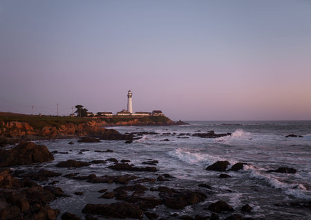 White lighthouse on the mountain coast of the ocean at sunset. Rocky beach with waves in the evening of summer against the background of a purple evening sky without cloudsの写真素材