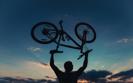 The athlete holds the bike on outstretched arms against the background of the evening blue summer sky and clouds. Young guy with a bicycle at sunset day after a bike rideの写真素材
