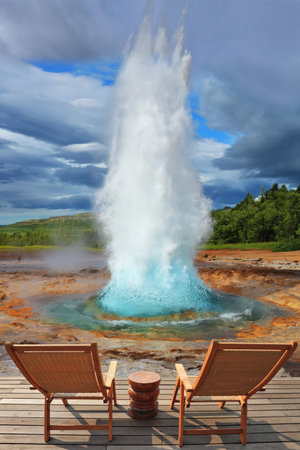 Pillar of hot water and steam from forcing its way out of the ground. Two lounge chairs and  small table on  wooden platform for easy observation.  Geyser Strokkur in Icelandの写真素材