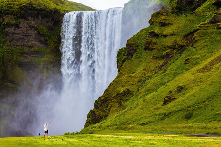 Grand waterfall Skogafoss in Iceland. Tourist in shirt and bandana threw up his hands with delight the beauty of nature Grand waterfall Skogafoss in Iceland. Tourist in shirt and bandana threw up his hands with delight the beauty of nature