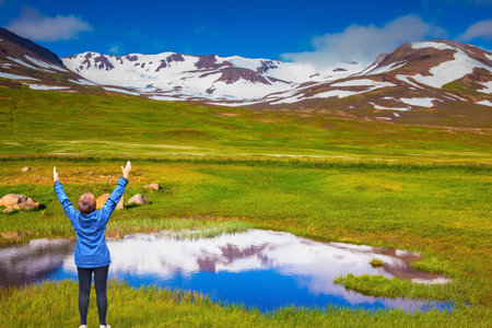 Blue lake water reflects the snowy hills. Iceland. The woman - tourist in blue jacket raised her hands in delight beauty of natureの写真素材