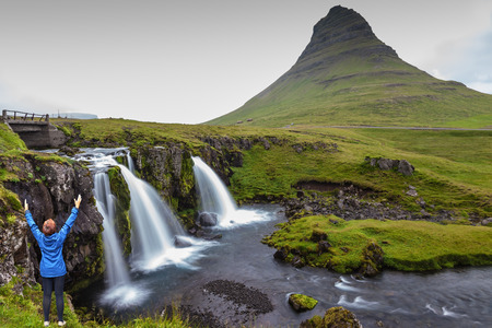 Threaded full-flowing waterfall Kirkjufell Foss on the grassy mountains. Middle-aged woman tourist admires the beauty of natureの写真素材