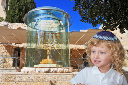 Cute little boy with long blond curls and blue eyes in a knitted skullcap. He stands at the Golden Menorah in Jerusalem. The Jewish holiday of Sukkotのeditorial素材