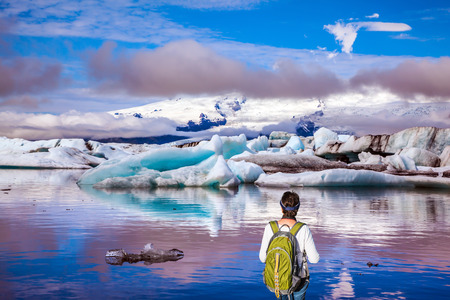 The woman - tourist with a green backpack watching the moving ice. Sunrise in the Ice Lagoon. Ice floes are reflected in the smooth water surface. The concept of extreme northern tourismの写真素材