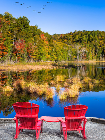 Red plastic loungers on the shore of the pond. Flock of migratory birds flies over the lake. Magnificent resort in French Canada - Mont Tremblant. Concept of ecological tourismの写真素材