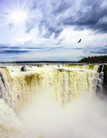 The autumn sun shines through the clouds. Andean condors fly above the roaring water. Devil's Throat is the part of the Iguazu Falls. Concept of active and photographic tourismの写真素材