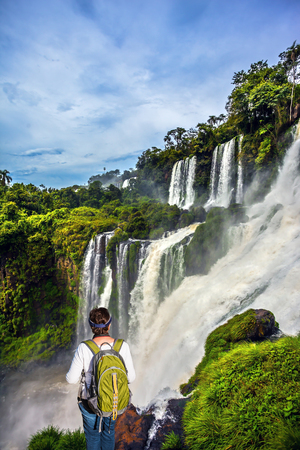 Energetic woman with a tourist backpack watching waterfalls. Scenic basaltic rock formations famous waterfalls Iguazu Falls. Concept of active and extreme tourismの写真素材