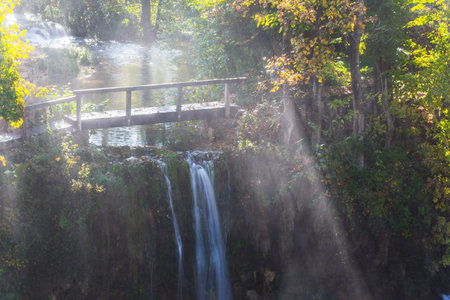 Sun rays over a wooden bridge and a waterfall. The small Croatian town of Slunj. Cascade of waterfalls on the Sluncica River. The concept of ecological, active and photo tourismの写真素材