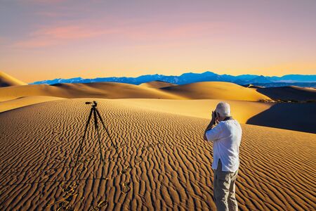 Sunset in desert. Gray-haired man - tourist in white T-shirt photographs magnificent landscape. Tripod standing by. Mesquite Flat Sand Dunes, Death Valley. Concept of active and photo tourismの写真素材