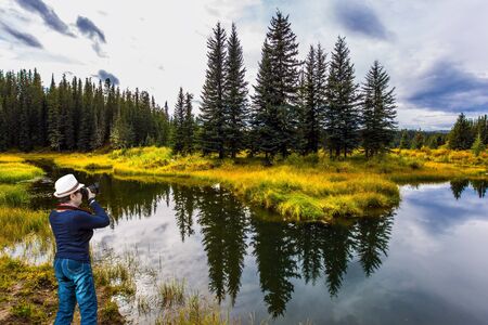 Northern Cordillera. Middle-aged woman in a beautiful hat takes pictures of the lake. Cold cloudy autumn day. The Northern Rocky Mountains. The concept of ecological, active and photo tourismの写真素材