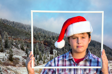 Charming boy in a clownish cap of Santa Claus smiles through a white frame. The first snow....