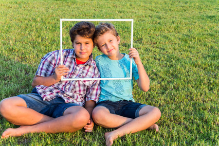 Background - green summer lawn. Concept - portrait and advertising photo. Two cute boys - brothers smiling, looking through a white frameの写真素材