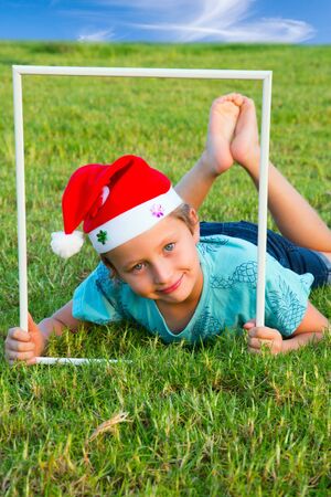 Charming boy in a clownish cap of Santa Claus smiles through a white frame. The background is green lawn. Concept portrait and advertising photoの写真素材