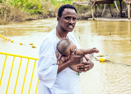 QASR El-YAHUD, ISRAEL - MARCH 2, 2020: The site of the baptism of Jesus Christ on Jordan River. Baby Baptism Ceremony. Father in a white shirt stands in the water and holds a baby. The concept of phototourismのeditorial素材