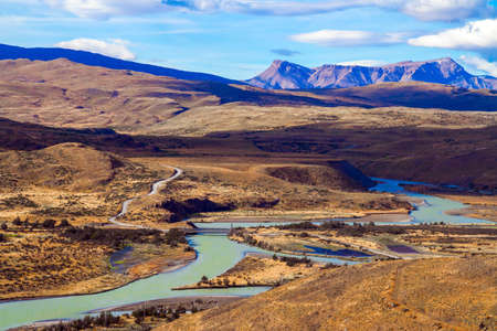 Shallow river with glacial melt water. Southern Hemisphere. Patagonia. Dirt road among yellowing grass. The magnificent park of Torres del Paine. The concept of extreme tourismの写真素材