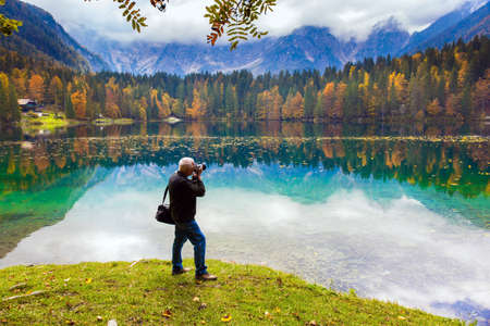 Man with a photo bag photographs a magnificent landscape. Alps. Lake Fuzine. Yellow trees are reflected in the green smooth water of the lake. Northern Italyの写真素材