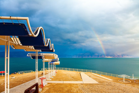 Picturesque beach with bright sunshades. The gloomy sky with dark thunderclouds and rainbow. The Dead Sea. Israeli coast. The exotic resort for treatment and relaxationの写真素材