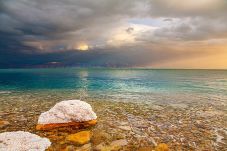 The sun illuminates the Jordanian coast. Israeli coast. Rocky beach covered with evaporated salt. The Dead Sea, sunset. Gloomy sky with dark thunderclouds.