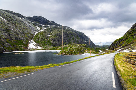 Narrow and dangerous mountain road glitters from the rain. Gray rocks surround the lake. The roadside is marked with high poles. Cold summer in Norway. Traveling by camper on road 520.の写真素材