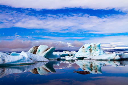 Clouds reflected in the smooth water of the largest glacial lagoon Jokulsaurloun. White and blue icebergs and ice floes. Iceland. Cold early summer morning. The concept of northern and photo tourismの写真素材