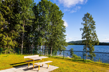 The shore of the lake is overgrown with grass and wildflowers. Warm July in Norway. Cozy corner for tourists. Picnic table and benches by the lakeの写真素材