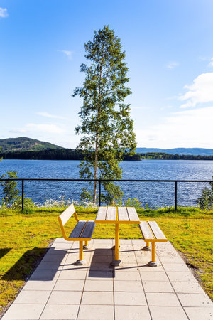 Warm July in Norway. Picnic table and benches by the lake. Cozy corner for tourists. Light white clouds in the blue sky illuminated by the sunrise. Magical summer morning.の写真素材