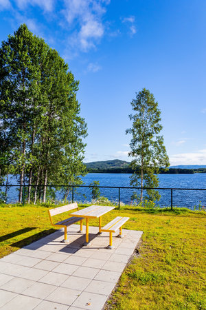 Picnic table and benches by the lake. Cozy corner for tourists. Magical summer morning. Light white clouds in the blue sky illuminated by the sunrise. Warm July in Norway.の写真素材