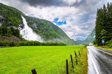 Roaring high-water cascading waterfall Vidfossen. Lush clouds and constant cold rain. Scenic road in the mountains of Western Norway. White water foam envelops the foot of the waterfallの写真素材