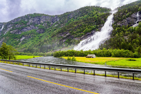 Huge powerful cascading waterfal Vidfossen. The rain-wet highway glitters in the fog. Western Norway. White water foam envelops the foot of the waterfall. Summerの写真素材