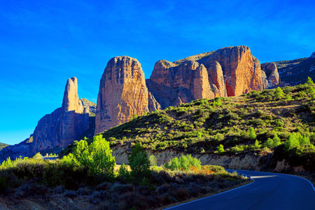 Asphalt highway passes through the rocks. The foothills of the Pyrenees. Sunny afternoon. The magnificent Mallets of Riglos is a conglomerate of rock formations. Romantic trip to Spainの写真素材