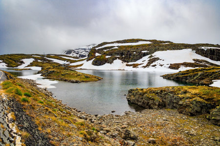 Gorgeous "Snow Road" in Norway. The cold water of the lake is covered with ice floes. Huge cold snowfields and sharp stone placers. Cold July in Northern Europe.の写真素材