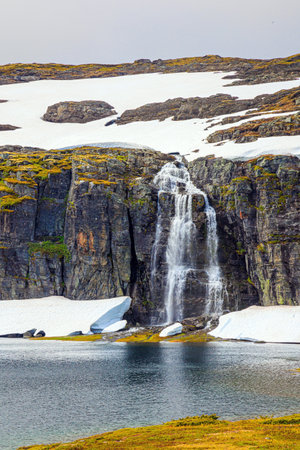 Tall waterfall falls into the lake. Gorgeous "Snow Road" in Norway. Incredible trip to the north of Norway in cold July. Mountains are covered with non-melting snowfieldsの写真素材