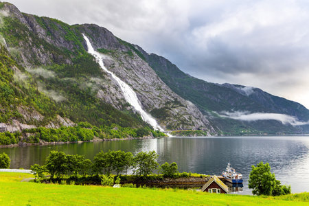 The giant scenic Langfossen waterfall is reflected in the smooth water of the Acre Fjord. Passes through the highway. Summer cold and rainy day. Western Norway.の写真素材