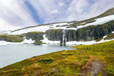 Tall Flotane waterfall falls into the lake. Snow road. Incredible trip to the north of Norway in cold July. TMountains and valleys are covered with non-melting snowfieldsの写真素材