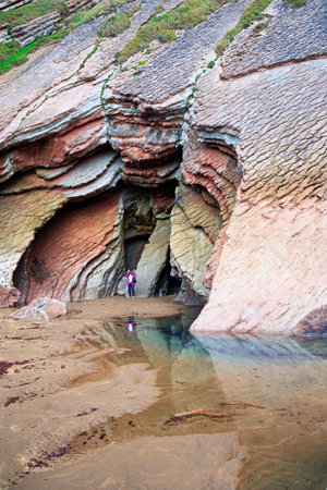Picturesque cave on the beach. Flysch - unique landscape of underwater rocks exposed at low tide. Woman in pink jacket photographs Playa Zumaya. Low tide in the Atlantic.の写真素材