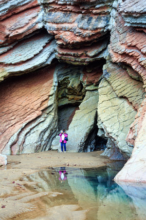 Picturesque huge cave on the beach. Woman in pink jacket photographs Playa Zumaya. Low tide in the Atlantic. Flysch - unique landscape. The Basque Country - Itsurun.の写真素材