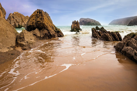 The smooth wet sand of the beach reflects the rocky boulders. The Playa de la Arnia beach. Snow-white sea foam glistens. Spain, Cantabria.の写真素材