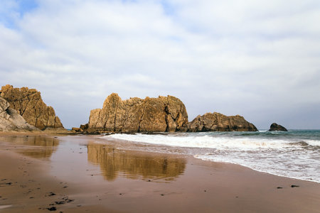 The smooth wet sand of the beach reflects the rocky boulders. The Playa de la Arnia beach.  Cantabria, Spain. Picturesque geological formations amaze tourists. Snow-white sea foam glistensの写真素材