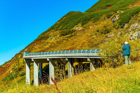 Gray-haired photographer in a jacket photographs the landscape. Austria. The most famous high mountain road is Grossglocknerstrasse. The bridge. Sunny day in October.の写真素材